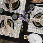 Overhead view of a set dining table with hand-printed placemats featuring gold stripes, folded Nandi Gold Napkins (Set of 4) by John Robshaw with napkin rings, plates, cutlery, glasses, a loaf of bread, flowers, and a covered dish. - 31263982714926