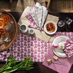 Table set for breakfast with John Robshaw's hand-printed, 100% cotton Sabah Metallic Napkins (Set of 4), a basket of pastries, a cup of coffee, a cinnamon roll on a plate, silverware, and a bouquet of flowers on a dark wooden table. - 31263994445870