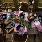 A rustic dining table is elegantly set with Nandi Berry Placemats by John Robshaw, featuring a charming pink hand block print. The setup includes black bowls and plates, stainless steel cutlery, freshly baked bread, aromatic candles, and an array of delectable foods such as asparagus and cheese. Natural light from a nearby window enhances the ambiance. The Nandi Berry Placemats are made in India from 100% cotton. - 31272680751150