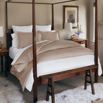 Under a dark wood canopy frame, a neatly made bed showcases the Velvet White Quilt from John Robshaw, complemented by white sheets. Matching velvet pillows and throws add a touch of elegance. In the background, a bedside table holds a lamp and a framed picture hangs on the wall. - 31258995818542