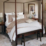 A neatly arranged bed featuring beige and white bedding, handcrafted with block prints from India, sits under a wooden canopy frame in a tastefully decorated bedroom. The room includes nightstands with lamps and artwork adorning the walls. Enhance the setting with the Sonal Decorative Pillow by John Robshaw, noting that pillow inserts are sold separately. - 31263563939886