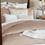 A neatly made bed with hand-quilted, beige patterned bedding and white pillows, accented by a floral decorative pillow. A nightstand with a green glass vase containing purple flowers is visible next to the bed, highlighting the exquisite craftsmanship of Indian artisans. Draped elegantly across the bed is the Velvet White Throw from John Robshaw. - 31258996604974