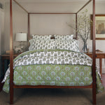 A wooden four-poster bed with patterned green and white bedding features a soft Jeeva Sage Quilt by John Robshaw, adorned with a delicate spring meadow design. Decorative pillows complement the scene, while a bedside table with a lamp on the left and a vase with branches on the right complete the cozy setup. - 31258910949422