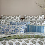 A neatly arranged bed with multiple pillows in various patterns and the John Robshaw Jeeva Sage Quilt, soft cotton featuring floral designs in blue, green, and white hues. Branches with small flowers are placed beside the bed. - 31258910982190