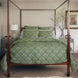A bedroom with a hand quilted four-poster bed featuring the John Robshaw Jeeva Sage Quilt, a white lamp on a nightstand, a framed picture on the wall, and a branch in a vase on the opposite nightstand.