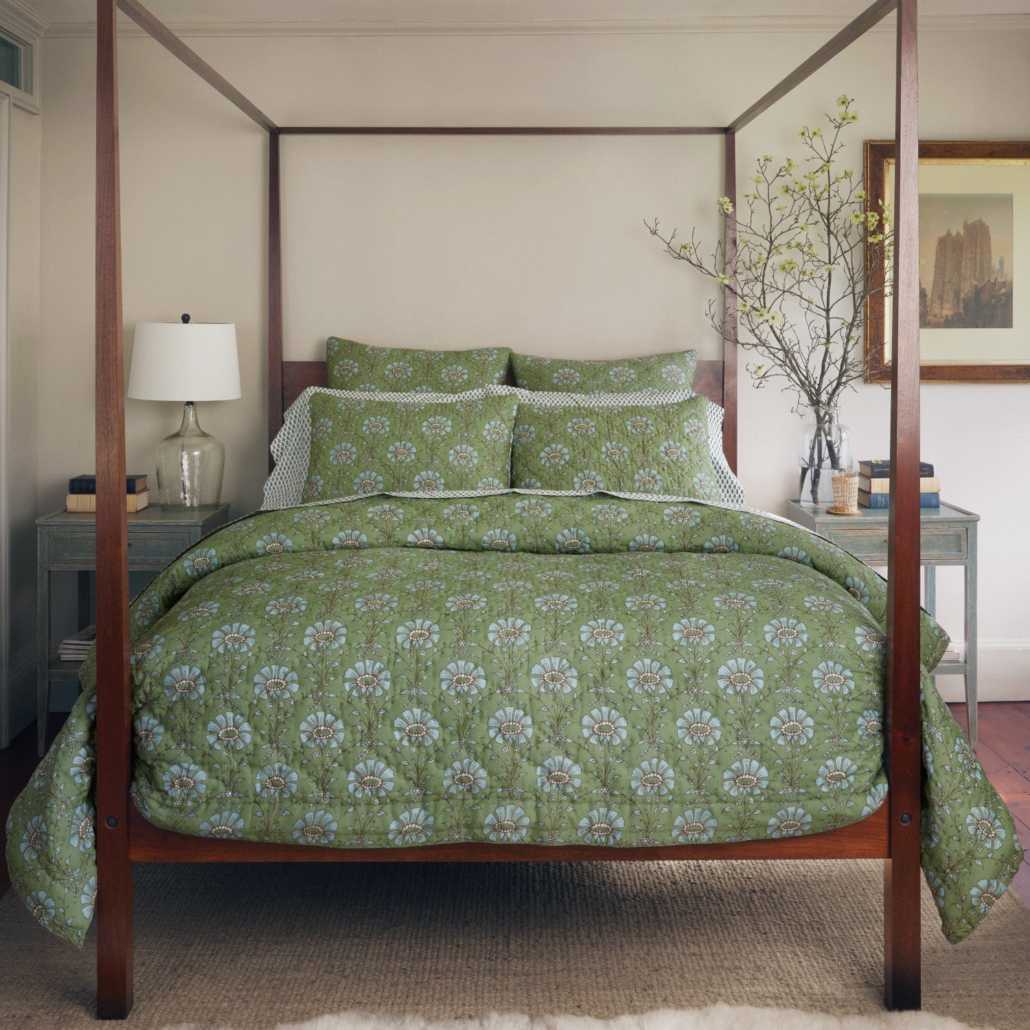 A bedroom with a hand quilted four-poster bed featuring the John Robshaw Jeeva Sage Quilt, a white lamp on a nightstand, a framed picture on the wall, and a branch in a vase on the opposite nightstand.