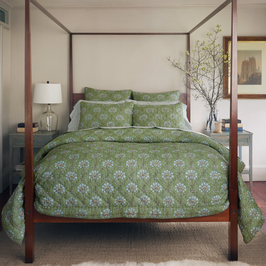 A bedroom with a hand quilted four-poster bed featuring the John Robshaw Jeeva Sage Quilt, a white lamp on a nightstand, a framed picture on the wall, and a branch in a vase on the opposite nightstand.