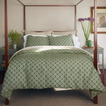 A neatly made bed with a green patterned comforter and cotton and linen pillows, flanked by bedside tables adorned with decorative items, a Woven Sand King Euro pillow from John Robshaw, and flowers in a vase. - 31263902924846