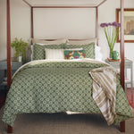 A neatly made bed with a green patterned duvet and pillows is flanked by bedside tables holding plants and decorative items. A striped blanket with a hand-sewn fringe is draped over the corner of the bed frame, and a Fringed White Bolster by John Robshaw adds a unique touch. - 31259445919790