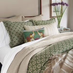 A neatly made bed showcases green patterned pillows and white linens topped with a beige quilt. Decorative upcycled linen pillows adorned with elephant designs and hidden zipper closures are artfully arranged, alongside a John Robshaw Fringed White Bolster. A vase of purple flowers adds a touch of charm to the bedside table. - 31259445952558