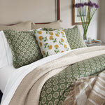 A neatly made bed featuring green and white patterned bedding, complemented by two green patterned pillows and one Woven Sand King Euro pillow by John Robshaw. A beige blanket with tassels is folded at the foot of the bed. On the nightstand sits a vase filled with purple flowers. - 31263902957614