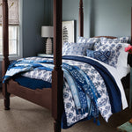 A neatly arranged four-poster bed, adorned with a variety of blue and white patterned pillows and blankets, features the Malvika Lapis Throw by John Robshaw in 100% hand-printed cotton voile. Tassels add a touch of elegance to the well-lit bedroom, where a window, lamp, and framed art are visible in the background. - 31259043069998
