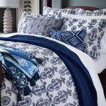 A neatly made bed adorned with blue and white floral-patterned bedding, accented by two Razia Lapis Decorative Pillows from John Robshaw, a dark blue quilt, and a folded blanket at the foot of the bed. The hand block printed designs on the bedding add a touch of elegance, showcasing craftsmanship made in India. - 31263385911342