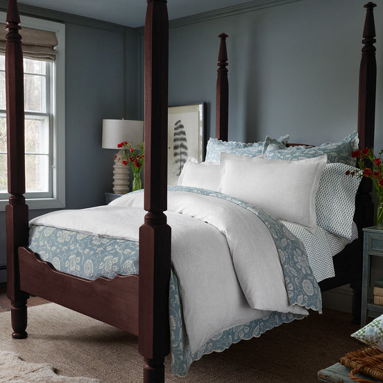 A sophisticated bedroom featuring a wooden four-poster bed, adorned with the John Robshaw Amina Ecru Duvet Set, crafted from the finest quality cotton linen. A nightstand with a lamp, flowers in a vase, and a window with light streaming in complement the light blue walls.