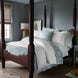 A bedroom featuring a dark wooden four-poster bed adorned with the luxurious Amina Ecru Duvet Set by John Robshaw, showcasing the finest quality cotton linen in white and blue hues. Nearby, a bedside table displays flowers alongside a lamp, while a window with blinds offers light control. The walls are painted in a serene muted blue-grey tone.
