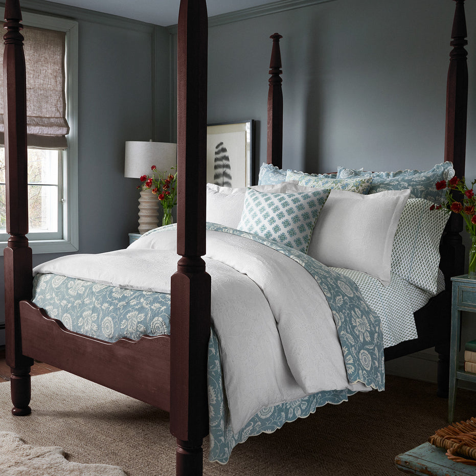 A bedroom featuring a dark wooden four-poster bed adorned with the luxurious Amina Ecru Duvet Set by John Robshaw, showcasing the finest quality cotton linen in white and blue hues. Nearby, a bedside table displays flowers alongside a lamp, while a window with blinds offers light control. The walls are painted in a serene muted blue-grey tone.
