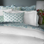 A neatly made bed adorned with the Kimari Mist Coverlet by John Robshaw, crafted in soft jacquard cotton chenille with intricate scalloped edges, complemented by multiple pillows in various shades of blue and green. Nearby stands a bedside table holding a stylish lamp and a vase filled with vibrant red flowers. - 31258913800238