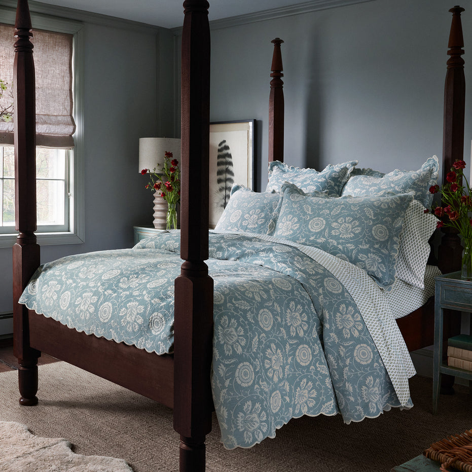 A bedroom featuring a four-poster bed adorned with the Kimari Mist Coverlet from John Robshaw, showcasing blue floral bedding made of soft jacquard cotton chenille. Bedside tables flank the bed, each holding a vase of flowers. In the background, a window decorated with a roman shade and framed artwork contribute to the room's delicate charm.