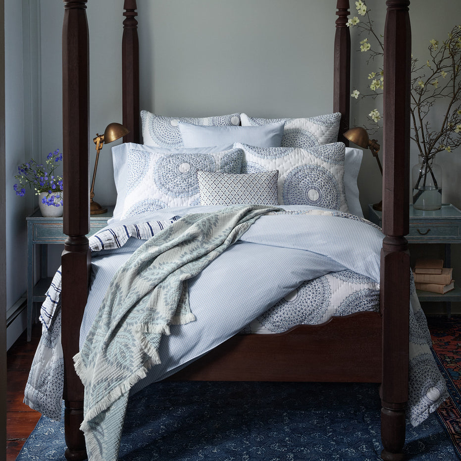 A wooden four-poster bed adorned with John Robshaw's Cinde Light Indigo Organic Duvet, patterned blue and white stylized print bedding, and pillows. It is situated in a room with light gray walls and complemented by a blue nightstand decorated with flowers and books.