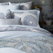 A neatly made bed displays patterned pillows, a quilt with blue and white designs, and the Cinde Light Indigo Organic Duvet by John Robshaw. Beside the bed sits a wooden nightstand adorned with a brass lamp and a vase of flowers.