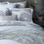 A neatly made bed displays patterned pillows, a quilt with blue and white designs, and the Cinde Light Indigo Organic Duvet by John Robshaw. Beside the bed sits a wooden nightstand adorned with a brass lamp and a vase of flowers. - 31249535664174