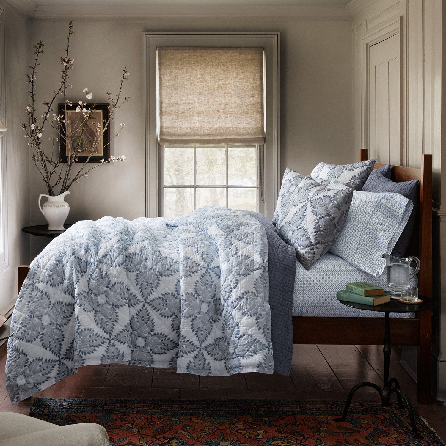 A wooden bed adorned with blue and white patterned bedding, featuring a Dasati Lapis Quilt by John Robshaw, stands next to a bedside table holding books and a glass of water. It is placed near a window with a beige shade, accompanied by a vase filled with branches.