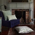 A cozy reading nook with a dark blue chair, patterned cushions featuring delicate hand-stitched edging, the Neyda Decorative Pillow by John Robshaw on the floor with a hidden zipper closure, a brick fireplace, and a lamp, all surrounded by rustic decor and greenery. - 31277085032494