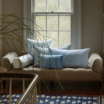 A beige couch sits in front of a window, adorned with several John Robshaw Palak Decorative Pillows featuring blue and white patterns. A plant branch extends over the back, and a blue and white hand block printed rug with hand-stitched edging is visible in the foreground. - 31263353831470