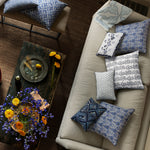 A beige couch is adorned with various blue and white patterned cushions, including the John Robshaw Kiara Decorative Pillow. A wooden coffee table holds a bouquet in a vase, yellow flowers, and a tray with glasses. - 31261336764462