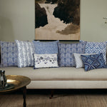 A sofa adorned with a selection of Sehra Azure Decorative Pillows by John Robshaw, showcasing various blue and white patterns including hand block printed designs, arranged before a wall painting of a waterfall. In the foreground sits a brass tray table. - 31263491555374