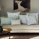 A beige sofa adorned with seven patterned pillows in various sizes and colors, including a John Robshaw Pipli Moss Bolster and one with hand-stitched edging, arranged on it. A painting of a waterfall graces the wall behind, while a small table with a black vase stands in the foreground. - 31263367036974