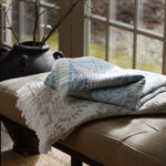 Two folded Dasati Sand throws by John Robshaw, featuring stonewashed cotton with intricate woven designs and fringed edging, are placed on a cushioned bench in front of a window, with a black vase in the background. - 31259045756974
