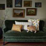 A dark green velvet sofa adorned with five assorted decorative pillows, including the elegant John Robshaw Kia Marigold Kidney Pillow, sits below various framed artwork on the wall. A potted plant is visible on the left side of the image. - 31259686469678