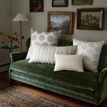 A green velvet sofa adorned with John Robshaw's Fringed White Bolster, multiple patterned cushions, framed artwork on the wall, a floor lamp, and a side table with a vase of flowers. - 31259446313006