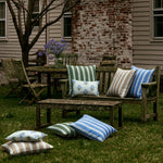 A Wooden outdoor furniture set with John Robshaw's Vintage Stripe Azure Outdoor Decorative Pillows placed on and around it, situated in a grassy backyard. An old brick chimney is seen in the background. Purple flowers adorn the table. The sun and rain resistant design ensures durability for all seasons. - 31272686452782