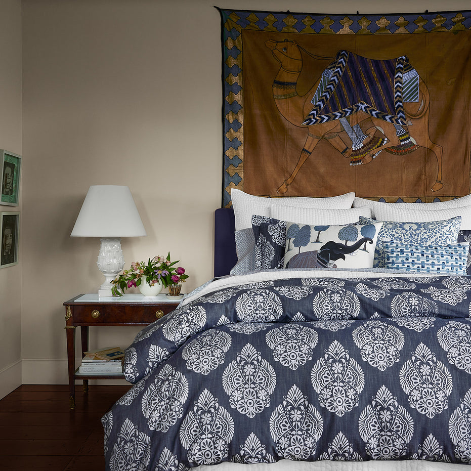 A hand-painted wall hanging complements the Indigo Elephant Decorative Pillow by John Robshaw on a bed, adorned with cotton linen.