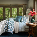 A bedroom with ivory bedding, a hand woven John Robshaw Chahan Ivory Throw, and flowers. - 30784360153134