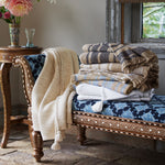 A stack of John Robshaw Ekram White Throws, a woven blend with fringed edges, on a bench in front of a window. - 30784359628846