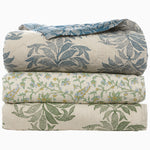 Three Asma Peacock Woven Quilts from John Robshaw, each featuring floral patterns, are stacked on top of each other. The top quilt is a cotton chambray design in blue and beige, the middle one is white with green and blue accents, and the bottom one is white with green patterns. Each quilt boasts a reversible print for added versatility. - 31274123558958