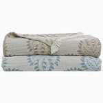 Two folded, patterned blankets stacked on top of each other; the top blanket, crafted from stonewashed cotton, features a beige and brown woven design, while the bottom blanket, the Dasati Lapis Throw by John Robshaw, is light blue with a blue and white design. Both blankets showcase delicate fringed edging for added texture. - 31274123165742