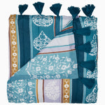 The Malvika Peacock Throw by John Robshaw is a folded, colorful blanket crafted from soft cotton voile, showcasing teal, white, and gold patterns with hand-printed designs and accented with teal tassels along one edge. - 31272464711726