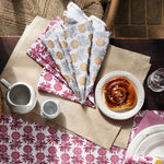 A table adorned with a 100% cotton, hand-printed floral tablecloth and placemats, set with a teacup, jug, glass of water, Sabah Metallic Napkins (Set of 4) by John Robshaw, and a plate holding a pastry. Made in India. - 31307875680302