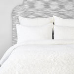 A white bed with a grey headboard and white pillows, featuring an Organic Hand Stitched Sand Quilt by Quilts & Coverlets. - 28271560523822