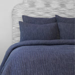 A bed with a hand-stitched, reversible Vivada Ink Woven Quilt made of super fine cotton chambray in blue. The bed also features a white headboard by John Robshaw. - 28271562358830