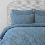 A reversible bed with a Vivada Peacock Woven Quilt made of cotton chambray coverlets and a white headboard by John Robshaw. - 28271561932846