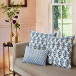 A John Robshaw Ojas Indigo Decorative Pillow rests on a couch in front of a pink wall, accompanied by a vase. - 30009690488878
