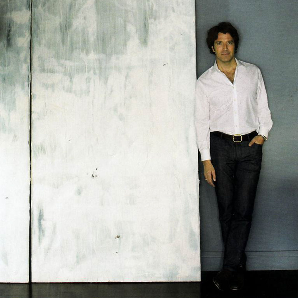 A man in a white shirt standing next to a white wall, showcasing Be Chalk White Among The Blank, an original work of art on canvas by John Robshaw.