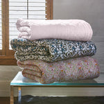 A Sagana Lotus Quilt and a reversible quilt from Quilts & Coverlets are stacked on top of a glass table. - 29553876729902