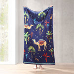 A woman is holding up a John Robshaw Dhule Indigo Beach Towel with camels and palm trees, perfect for Kenya safaris. - 29274375159854