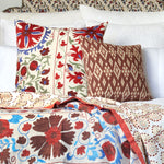 A bed adorned with a Red Suzani Mmm coverlet accented by embroidered tribal textiles from Central Asia, designed by John Robshaw. - 30264133713966
